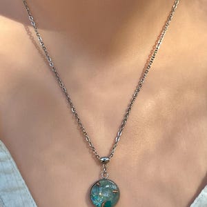 May include: A silver-toned necklace with a round pendant. The pendant features a teal and blue design with copper accents. The necklace is shown against a light background.