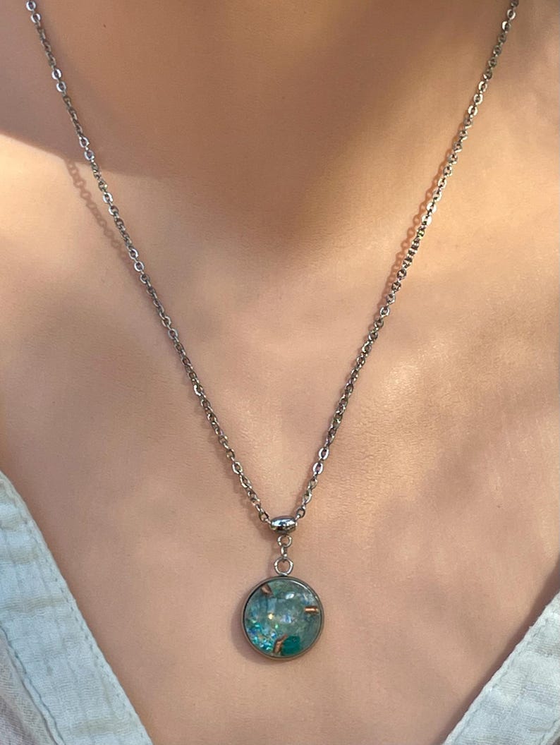 May include: A silver-toned necklace with a round pendant. The pendant features a teal and blue design with copper accents. The necklace is shown against a light background.