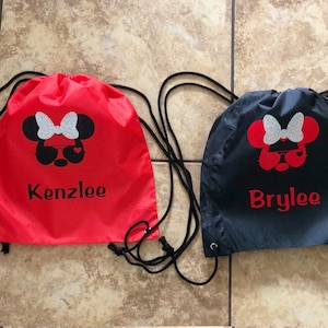 May include: Two red and black drawstring bags with a Minnie Mouse design and personalized names. The red bag says "Kenzlee" and the black bag says "Brylee".