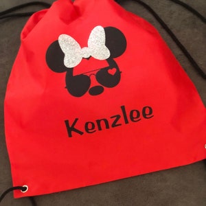 May include: Red drawstring bag with a black and white Minnie Mouse design with a silver glitter bow. The bag has the name "Kenzlee" printed in black letters.