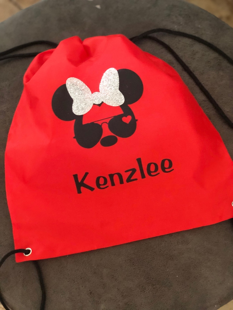 May include: Red drawstring bag with a black and white Minnie Mouse design with a silver glitter bow. The bag has the name "Kenzlee" printed in black letters.