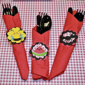 May include: Three red napkins with black plastic cutlery wrapped inside. Each napkin has a different paper cutout design: a bee, a watermelon, and an ant.