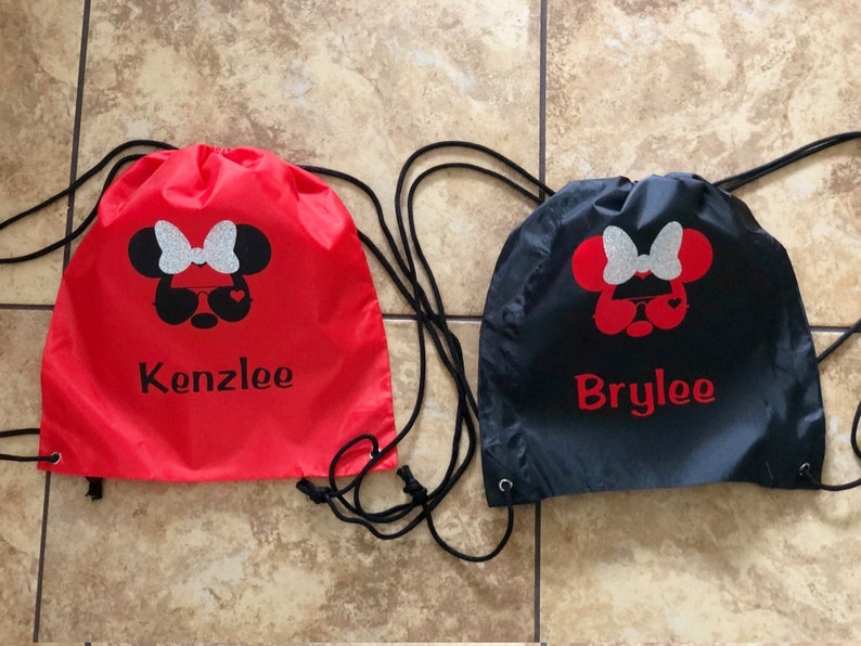 May include: Two red and black drawstring bags with a Minnie Mouse design and personalized names. The red bag says "Kenzlee" and the black bag says "Brylee".