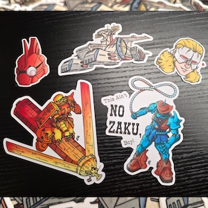 Puede incluir: Una colección de pegatinas coloridas con ilustraciones de dibujos animados de robots, una nave espacial y un personaje enmascarado. Una pegatina tiene el texto "This Ain't NO ZAKU, Boy!"