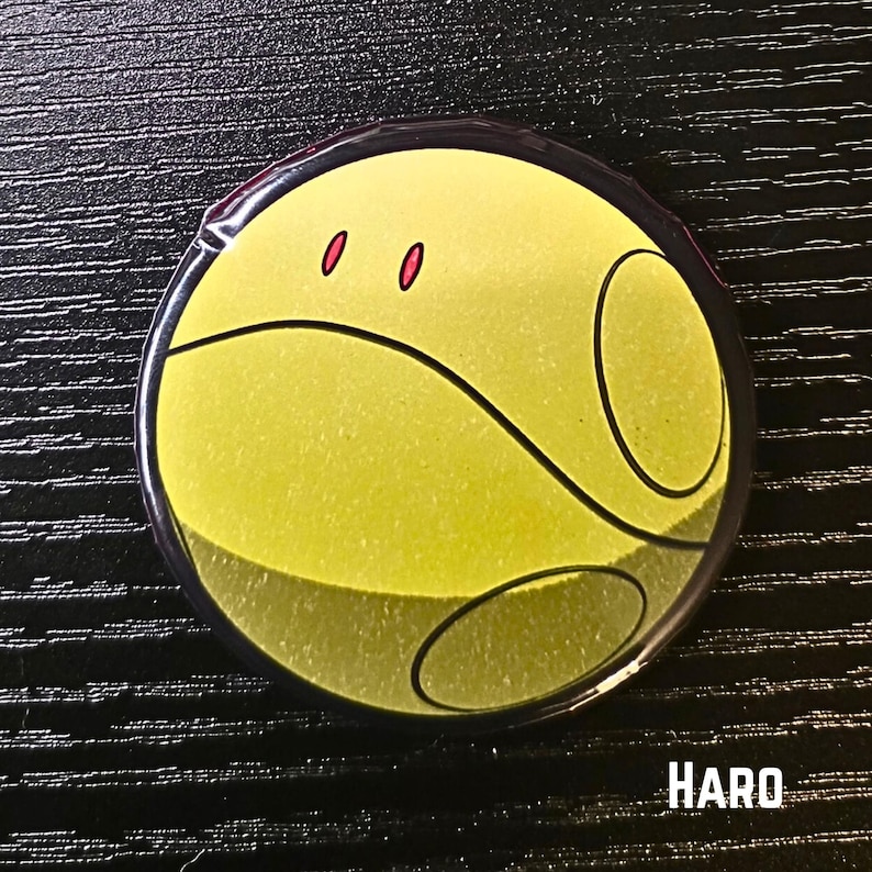 Puede incluir: Un bot&oacute;n redondo con una cara amarilla, l&iacute;neas negras curvas y dos ojos rojos. El bot&oacute;n tiene un borde negro y la palabra "HARO" en texto blanco en la parte inferior. Sobre un fondo negro.