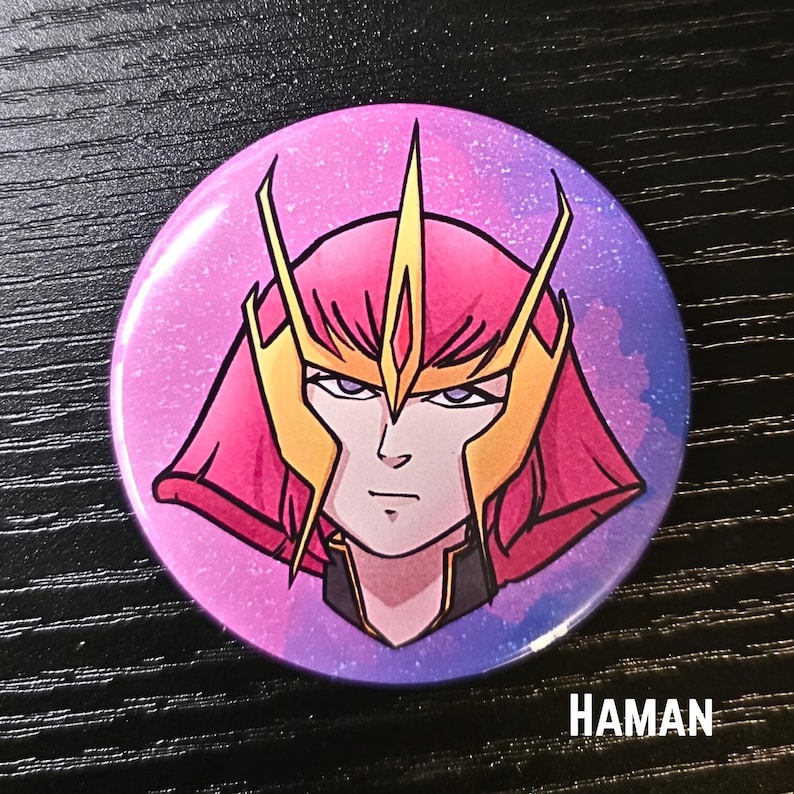 Puede incluir: Un bot&oacute;n redondo con una ilustraci&oacute;n de un personaje de anime. El personaje tiene cabello rosado y un tocado dorado, sobre un fondo degradado rosa y azul. La palabra "HAMAN" est&aacute; impresa en blanco.
