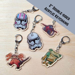May include: Collection of 2-inch double-sided acrylic keychains. The keychains feature various cartoon designs, including a stormtrooper helmet, a red and gray vehicle, a frog, and a lizard-like creature. The keychains have silver-colored metal clasps.