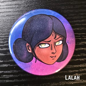 Puede incluir: Bot&oacute;n redondo con una ilustraci&oacute;n de dibujos animados de la cara de una mujer. La mujer tiene el pelo oscuro, ojos amarillos y un bindi rojo. El bot&oacute;n tiene un fondo de galaxia azul y p&uacute;rpura. El nombre "LALAH" est&aacute; impreso en la parte inferior.