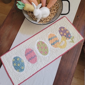 Peut inclure: Un chemin de table blanc avec une bordure rose présente cinq œufs de Pâques colorés. Les œufs sont décorés de motifs et de couleurs différents, notamment du bleu, du vert, du jaune et du violet. Un lapin en céramique blanc est assis dans un panier de carottes.