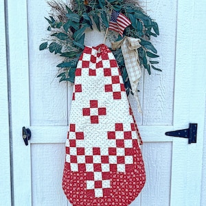 May include: A patriotic wreath with greenery, an American flag, and a plaid bow hangs on a white door. A red and white quilt with a checkerboard pattern is draped below the wreath, creating a festive display.