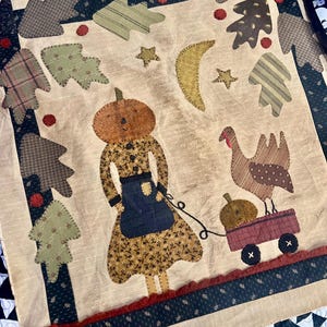 May include: A handmade quilt with a beige background featuring appliqued figures. A pumpkin-headed person pulls a wagon with a pumpkin and turkey. Trees, stars, and a crescent moon are also appliqued. The quilt has a dark blue border.