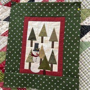 May include: A green fabric panel with a red border and a white background featuring a snowman and several green trees. The trees are made of different fabrics and have a brown trunk. The snowman is wearing a black top hat and a red scarf.