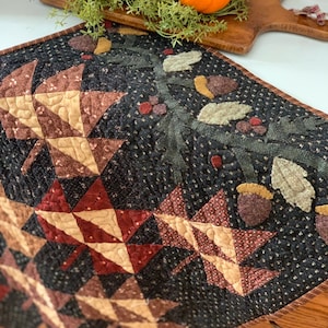 May include: A black quilted table runner with a fall theme. The runner features brown and tan leaves, acorns, and a black background with white speckles.