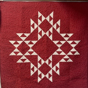 Może przedstawiać: Czerwono-biała kołdra z wzorem gwiazdy. Gwiazda składa się z białych trójkątów na teksturowanym czerwonym tle. Kołdra jest kwadratowa i wydaje się być wykonana ręcznie, ze szczegółowym wzorem ściegów.