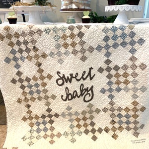 May include: A white quilt with a brown "sweet baby" inscription in the center. The quilt is made of squares in various shades of brown, blue, and white. The quilt is displayed on a table with a dessert spread and other baby shower decorations.
