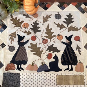 Peut inclure: Une tenture murale en tissu carré présentant une scène en appliqué de deux personnages en chapeaux et robes noirs près d'une vigne centrale avec des feuilles de chêne et des citrouilles. Un chat noir est assis entre eux, bordé de carrés de tissu crème.