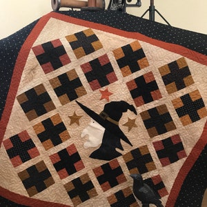 May include: A black and white quilt with a witch and stars design. The quilt is made with a variety of fabrics, including black, white, brown, and orange. The quilt is displayed on a black surface with a black crow figurine.