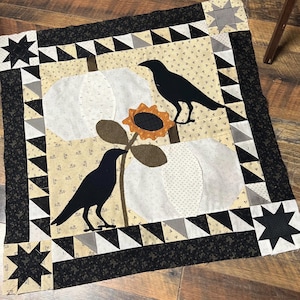 May include: A black and white quilt with a black and white star design in each corner. The center of the quilt features a brown stem with a large orange sunflower and two black birds. The background is a cream color with a small white polka dot pattern.
