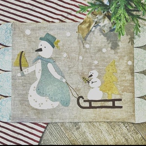 May include: A beige fabric wall hanging with a blue and white snowman pulling a smaller snowman on a sled. The background is a snowy scene with a green pine tree. The fabric has a scalloped edge.