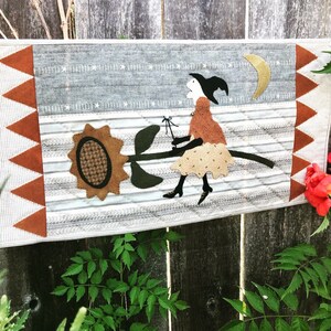 May include: A handmade wall hanging featuring a witch flying on a broomstick with a sunflower in the background. The witch is wearing a brown dress and a black hat. The background is a gray and white striped fabric with a crescent moon in the upper right corner.