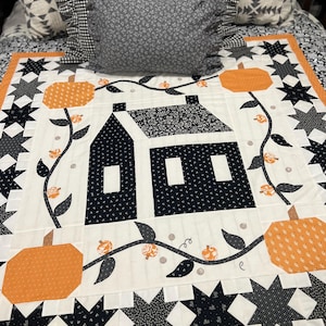 May include: A handmade quilt featuring a central house design in black and white, surrounded by orange pumpkins and star patterns. The quilt has an orange border and is set against a backdrop of patterned pillows.