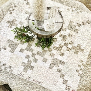 May include: A white quilted table runner with a gray checkered pattern and the text "let it snow" embroidered in the bottom left corner. A silver cake stand with a white ceramic house and two white ceramic trees sits on top of the runner.