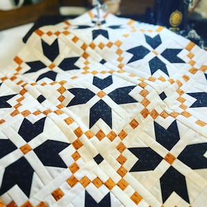 Może przedstawiać: Kołdra z geometrycznym wzorem czarnych, białych i pomarańczowych gwiazd. Kołdra jest szyta na maszynie do szycia.