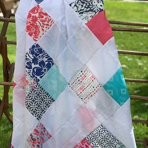 Könnte beinhalten: Eine weiße Steppdecke mit Rautenmuster und bunten quadratischen Flicken. Die Flicken zeigen florale, geometrische und abstrakte Designs in Rot-, Blau-, Rosa- und Türkistönen. Die Steppdecke ist über einer Holzkonstruktion drapiert.