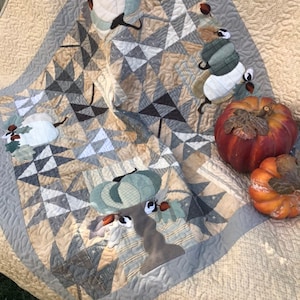 May include: A beige and grey patchwork quilt with an autumn theme. The quilt features a variety of fabrics, including plaid, stripes, and solids. The quilt is decorated with appliquéd pumpkins and leaves.