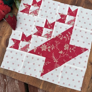 May include: A patchwork quilt block with a star pattern. The stars are red with a floral design, set against a white background with small red floral accents. The quilt block is on a wooden surface.