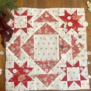 May include: A handmade quilt block with a patchwork design featuring red and cream floral and star patterns. The block is adorned with various vintage buttons. The quilt block is a square shape and is displayed on a wooden surface.