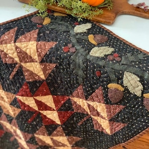 Pode incluir: Um corredor de mesa acolchoado com fundo preto e um desenho de folhas de outono em tons de marrom, vermelho e creme. As folhas são cercadas por uma borda de bolotas e folhas verdes.