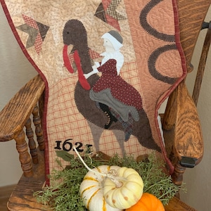 May include: A quilted wall hanging with a brown turkey and a woman in a red dress riding on its back. The quilt is decorated with stars and the number 1621. The quilt is hanging on a wooden chair with pumpkins in front of it.