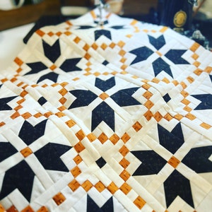 May include: A quilt with a geometric pattern of black, white, and orange stars. The quilt is being sewn on a sewing machine.