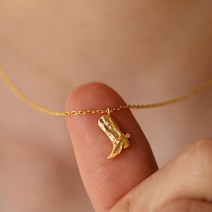 Peut inclure: Un collier en chaîne dorée avec un petit pendentif en forme de botte dorée. Le collier est porté par une personne.