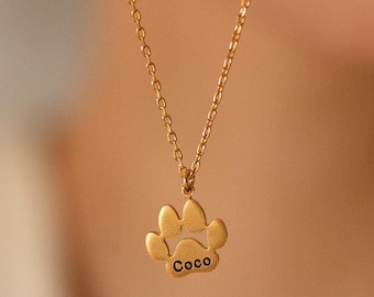 Puede incluir: Un collar de cadena dorada con un colgante con forma de huella de pata. El colgante tiene el nombre "Coco" grabado.