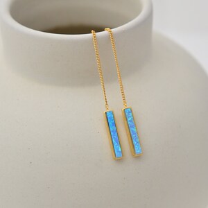 May include: A pair of gold-tone threader earrings featuring long, slender rectangular bars inlaid with vibrant, iridescent blue opal-like stones. The earrings consist of delicate gold chains that connect to the vertical bar pendants.