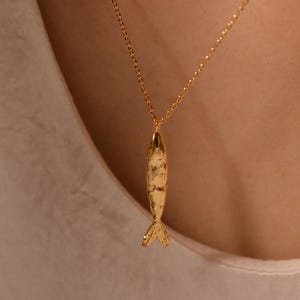 Puede incluir: Un collar con colgante de pez dorado. El colgante tiene una superficie texturizada y un diseño de aleta caudal. La cadena del collar es delicada y también dorada. El fondo es una persona con una camisa blanca.