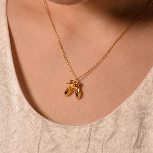 May include: A gold necklace with a pendant of two ballet slippers. The pendant hangs from a delicate chain. The necklace is worn against a cream-coloured top.