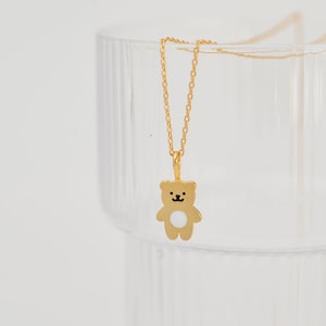 Könnte beinhalten: Eine goldfarbene Halskette mit einem kleinen bärenförmigen Anhänger. Der Bär hat eine glatte Goldoberfläche mit einem weißen kreisförmigen Akzent auf dem Bauch und schwarzen Emaille-Details für Augen, Nase und Mund, an einer feinen Goldkette.