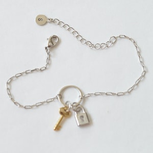 Può includere: Un braccialetto a catena d'argento con un piccolo ciondolo a forma di chiave e lucchetto. La catena ha una chiusura a moschettone e un piccolo tag d'argento con la lettera "S" su di esso.