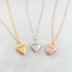 May include: Three heart-shaped lockets on delicate chains. One is gold with "HARPER" engraved, another is silver with "EMMA", and the third is rose gold with "LUCAS". The lockets are displayed against a soft, neutral background.