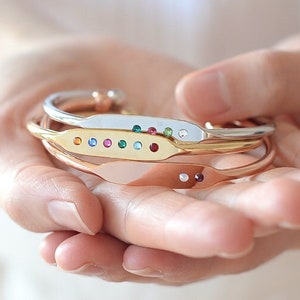 May include: Three metal bracelets in gold, silver, and rose gold are held in open hands. Each bracelet features a slender, curved design with a row of colorful gemstones. The bracelets are a delicate and elegant accessory.