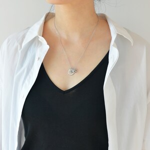 May include: A silver necklace with a small round pendant. The pendant has a textured design and is worn by a person wearing a white shirt and a black top.