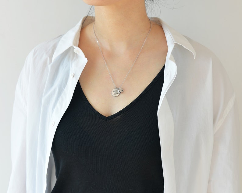 May include: A silver necklace with a small round pendant. The pendant has a textured design and is worn by a person wearing a white shirt and a black top.