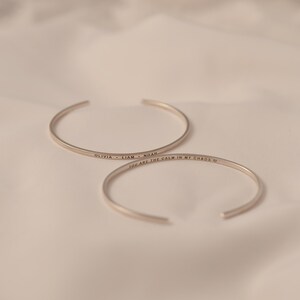 May include: Two silver-colored open cuff bracelets. One bracelet is inscribed with the names "OLIVIA &bull; LIAM &bull; NOAH". The other bracelet is inscribed with the phrase "YOU ARE THE CALM IN MY CHAOS" and a heart symbol.