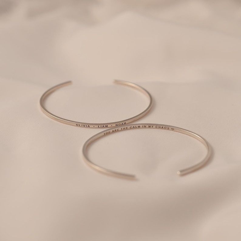 May include: Two silver-colored open cuff bracelets. One bracelet is inscribed with the names "OLIVIA &bull; LIAM &bull; NOAH". The other bracelet is inscribed with the phrase "YOU ARE THE CALM IN MY CHAOS" and a heart symbol.