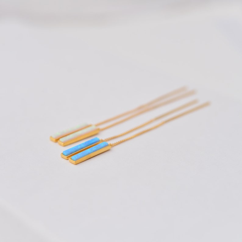 May include: A pair of gold-tone threader earrings featuring rectangular bar pendants. One earring has a bright blue inlay, while the other has a pale, iridescent white inlay. Each pendant is attached to a long, thin gold-tone chain.