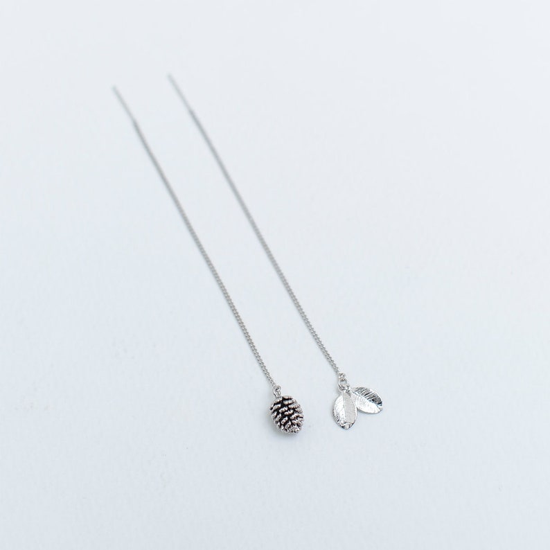 May include: A pair of silver dangle earrings with a pine cone charm on one earring and two leaf charms on the other.