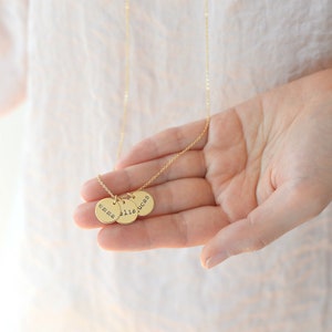 Op de afbeelding: Gouden ketting met drie ronde bedels, elk gegraveerd met een naam: "emma", "ellie" en "lucas".
